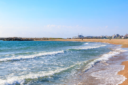Serene sandy beach with gentle waves and distant cityscape under clear blue sky - Powered by Adobe
