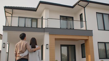 Rear view of a happy Asian couple embracing outside a modern white house, symbolizing new home ownership and real estate investment success.
