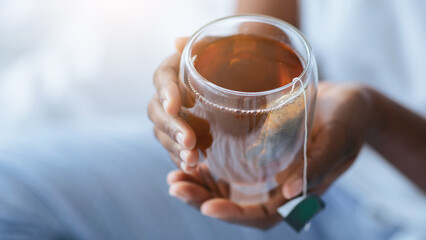 A person is holding a glass cup filled with tea. The cup has a tea bag attached by a string. The...