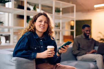 Pensive businesswoman drinking coffee while using cell phone in office.