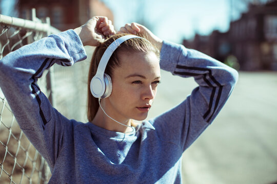 Young adult woman focused, tying hair with headphones on urban street