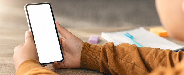 A young person sits at a wooden table holding a smartphone with a blank screen. Nearby are a...