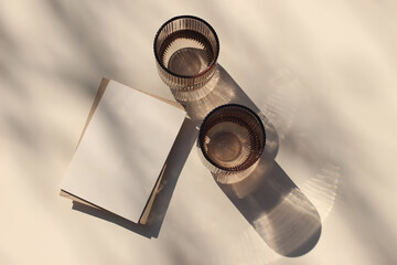 Blank greeting card, invitation mockup on beige table background. Two colored water glasses, bathed in sunlight casting shadows of leaves. Summer stationery, refreshing concept. Flat lay, top view