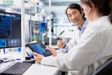 Smiling scientists in biotechnology laboratory looking at 3D molecular model on tablet. Happy researchers use bioengineering app on device in medical lab displaying DNA data