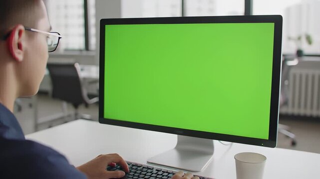 Young professional man typing on a green screen computer monitor in a bright modern office with copy space for digital content placement