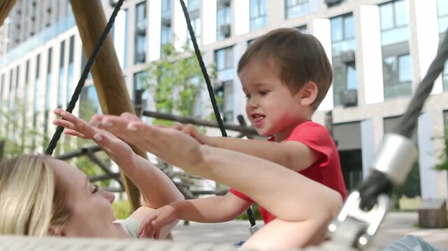 Mother having fun with her little son during a walk. Young mom plays patty-cake and swings on a swing with her baby