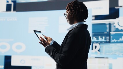 Manager using tablet to compare company metrics on digital videowall before making decisions. Entrepreneur analyzing key performance indicators using device app to optimize profitability, camera B