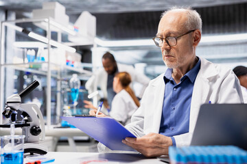 Elderly man in laboratory writing observation notes on clipboard document during experiment. Senior researcher recording test findings on paper to ensure data accuracy, doing reporting