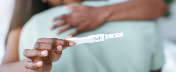 A couple embraces in a living room while one partner holds a pregnancy test. The mood is filled with anticipation and excitement in this intimate moment.
