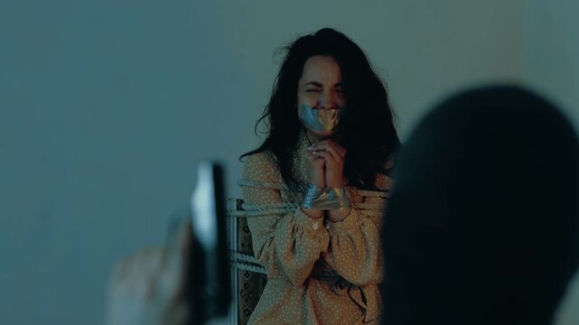 Man in a black balaclava threatening a woman with a gun. Lady tied with a rope, adhesive tape on her mouth and hands. Kidnaping, crime, violence concept. Slow motion