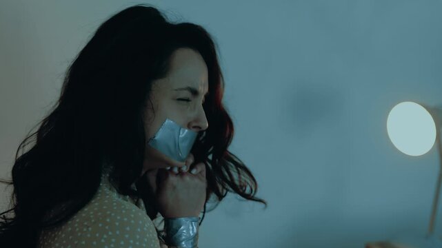 Abducted young woman sitting with her mouth sealed with tape, tied with a rope. Female hostage closing her eyes and praying for salvation. Kidnaping, crime, violence concept. Slow motion