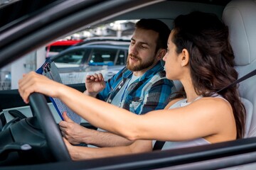 Young attractive woman driving car with male instructor in passenger seat during drive lesson. Man holding checklist. Driving test, driver courses, exam concept