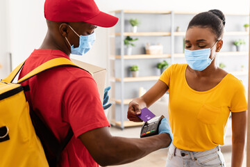 Obraz premium African American Client Lady Paying Courier For Delivery Of Cardboard Parcel Box Putting Credit Card To Terminal Machine Standing Indoors, Wearing Face Mask. Payment For Delivery Service Concept