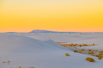 White gypsum dunes roll gently across the desert floor with distant mountains silhouetted beneath a warm sunset sky.