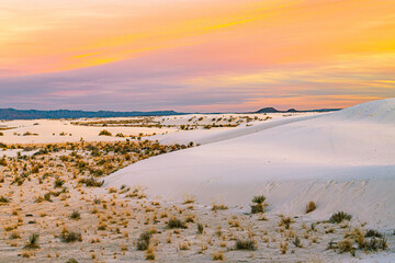Rolling white sand dunes dotted with desert vegetation glow under a colorful sunset sky at White Sands National Park in New Mexico. © ZL Visuals