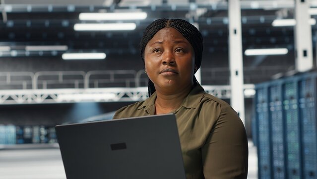 Technician in server room using laptop to upgrade rigs for increase in clients workload capacity. African american woman in data center configures equipment, improving processing speed, camera B - Powered by Adobe