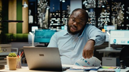 Male business director answers incoming phone call late at night, using laptop to read briefing data and engage in a transparent dialogue. Exchanges ideas to strengthen relations.