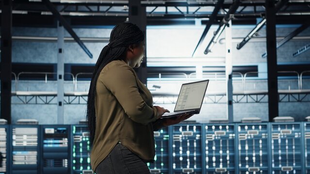 Software developer in data center using laptop to coordinate deployment of new server architecture. Server hub IT professional using notebook to do rollout of cybersecurity framework, camera B - Powered by Adobe