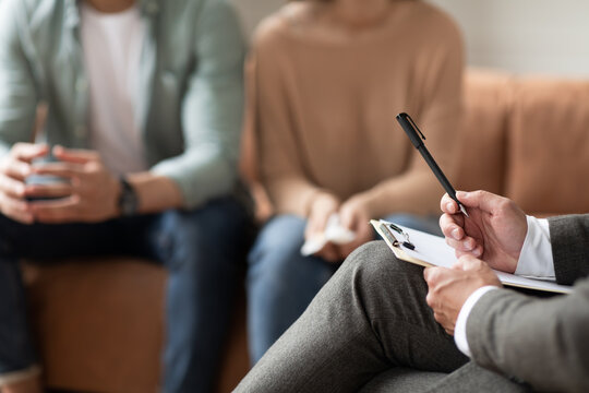 Unrecognizable couple having therapy session at psychotherapists office, selective focus on male professional psychologist holding pen and clipboard, writing in notepad, talking and consulting clients