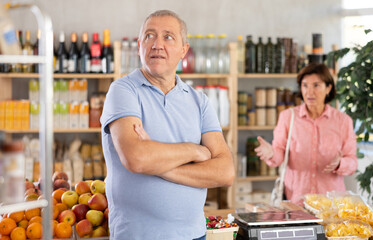 Senior wife stands behind pensioner husband and yells at companion. Angry man stand in front, he is...