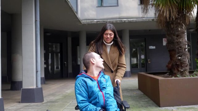 Young smiling caregiver happily pushing a man with a disability in a wheelchair through an urban courtyard, sharing a moment of connection and support while enjoying a pleasant walk together