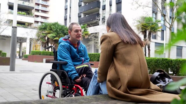 Young man with a physical disability sitting in a wheelchair having an animated conversation with a female friend or caregiver in a modern city park, enjoying a moment of connection and support