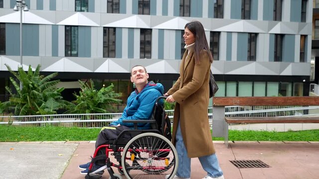 Young caregiver woman pushing a smiling man with a physical disability in a wheelchair through an urban park, representing support, accessibility, and positive inclusive relationships
