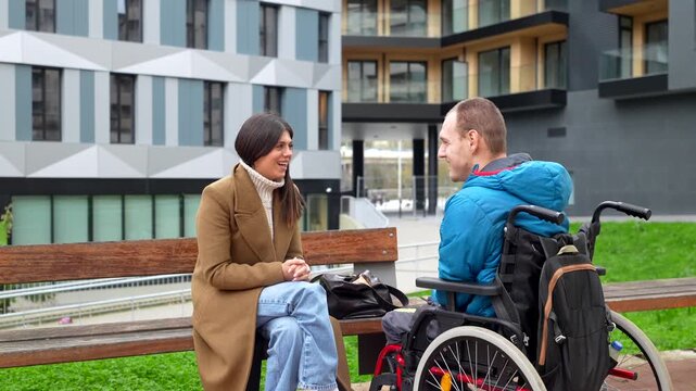 Supportive female social worker having a pleasant conversation with a young man with a physical disability in a wheelchair, sitting together on a bench in a modern city courtyard