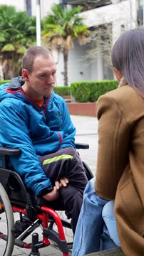 Young man with a physical disability sitting in a wheelchair, engaging in an expressive conversation and smiling with a woman in an urban park, showcasing friendship and social interaction