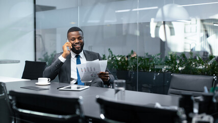 A man sits at a conference table in an office. He holds papers in one hand and talks on the phone...