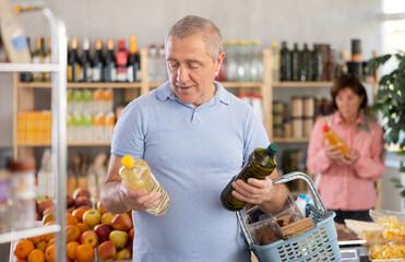 Mature man buyer looks thoughtfully at bottles of vegetable oil, woman client in background. Client examines product in detail, get acquainted with information for consumer