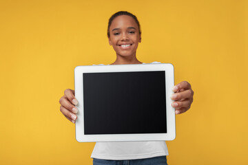 Smiling teen african american girl show tablet with empty screen, isolated on yellow background....
