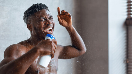A man is standing in a shower, holding a bottle and singing. Water is falling on him as he expresses joy. The setting is a bathroom with natural light coming through a window.