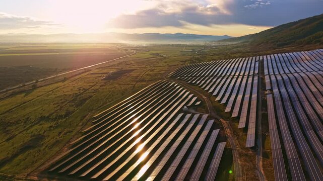 Rows of solar panels stretch across open land near mountains as the sun sets in the background. A winding road runs through the scene, with sunlight reflecting off the panels.