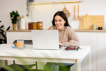 Young woman studying on a laptop while enjoying a cozy breakfast of coffee and croissants in her bright, modern kitchen. Education and e-learning concept