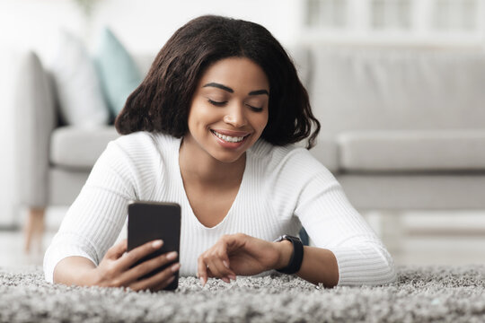 Happy african american woman using smartwatch with e-mail notifier and smartphone, resting at home in living room. Modern hand device notifying internet message - Powered by Adobe