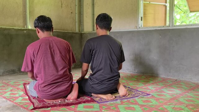 Two young Muslim men performing Salah prayer inside a wooden hut on a plantation. Showing prostrating (Sujud) and sitting (Tahiyat) positions. Peaceful Islamic spiritual activity in a rural area
