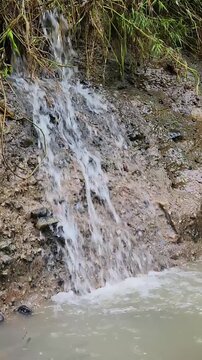 Small muddy waterfall pouring from eroded bank into cloudy pool. Rain runoff cascade dropping over wet soil cliff beside wild grass. Mini waterfall stream flowing down muddy slope into murky water.