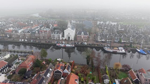 Aerial view of foggy Dutch canal town gabled brick homes, docked boats, and tall church tower form a tranquil, symmetrical waterfront scene softened by mist.