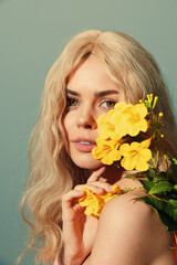 Beautiful young woman with long blonde hair holding yellow flowers against a soft blue background, exuding a serene and joyful emotion during springtime.
