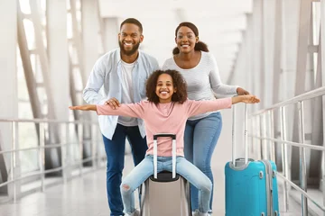 Fotobehang Muziek Happy Loving Family In Airport. Portrait of joyful excited black girl sitting on suitcase and laughing, spreading arms imitating plane, smiling cheerful parents posing, standing in terminal hall  © Prostock-studio
