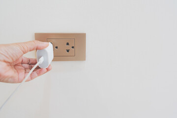 close up hand plugging adapter charge the battery with outlet socket on white concrete wall , for electric safety concept