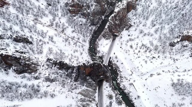 A dramatic top-down aerial view of a bridge, winding river and snowy canyon walls in Munzur Valley. Ideal for nature, winter landscapes, geography visuals and cinematic drone footage.