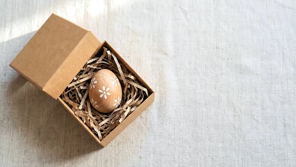 A brown egg with a white flower design is nestled in a bed of straw inside an open brown box on a white textured surface