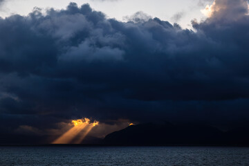 Early morning rays of sun strike through the dramatic clouds over the mountains of the Alaskan coast.