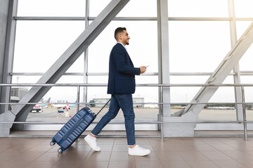 Young middle-eastern businessman with suitcase and smartphone in hands walking in airport terminal, handsome arab male going to flight boarding, ready for air trip, side view with copy space
