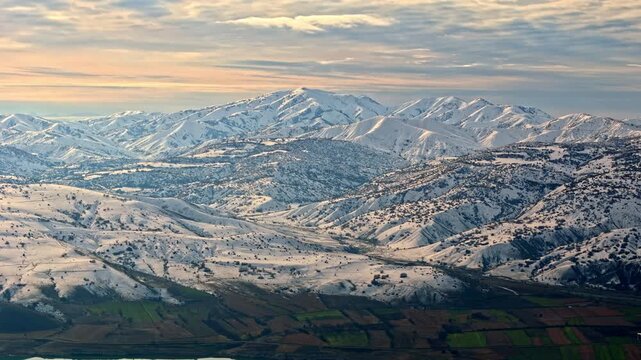 Aerial footage of a snowy mountain range and a wide winter valley with patchy farmland. Ideal for nature, winter landscapes, geography visuals, travel content and cinematic drone footage.