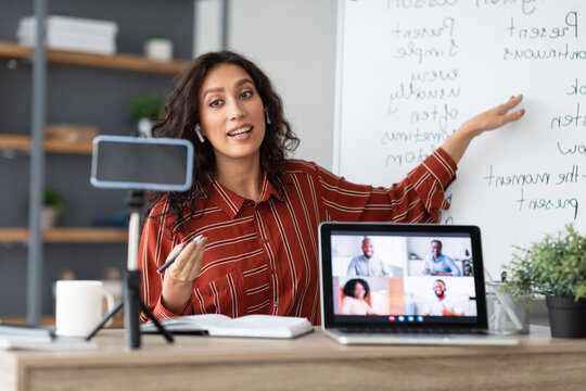 E-learning Concept. Portrait of young female teacher pointing at whiteboard, explaining English grammar rules to adult students. Female tutor recording lesson using smartphone on tripod