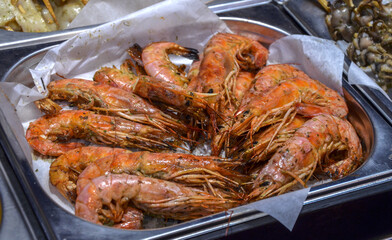 Metal tray holds a serving of large, whole, grilled prawns with their shells and heads intact, resting on white parchment paper