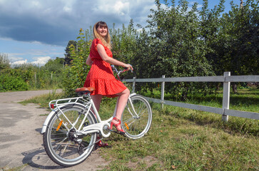 Woman in a red polka-dot dress rides a white bicycle in a sunny countryside setting. She smiles beside a wooden fence, embraced by green trees and open sky, exuding carefree leisure and fashion